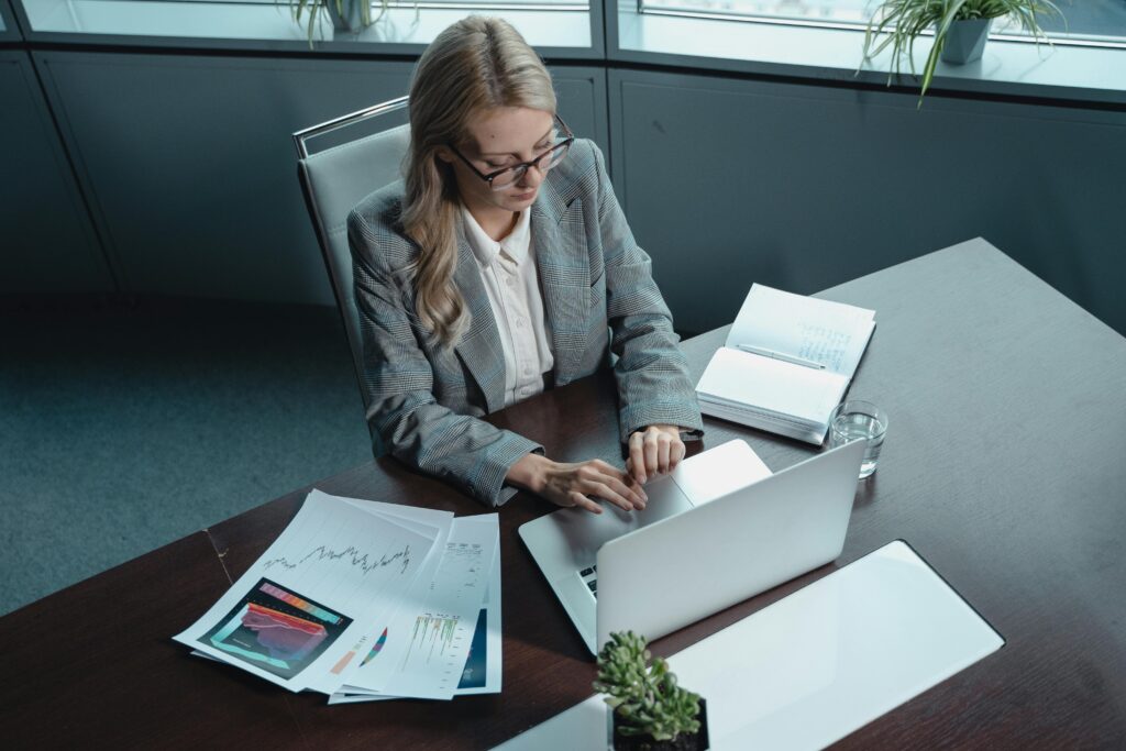 Blonde woman in a blazer working on a laptop in a modern office with documents.